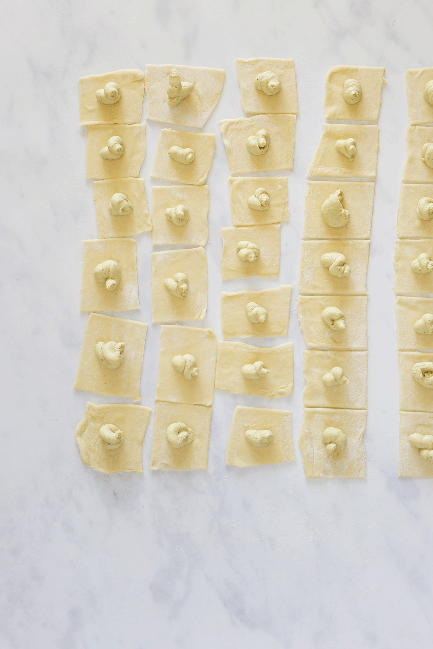 Squares of pasta dough laid out evenly on a white marble countertop. The image shows an animation that flashes back and forth between plain pasta dough squares and squares with a dollop of vegan ricotta in the middle.