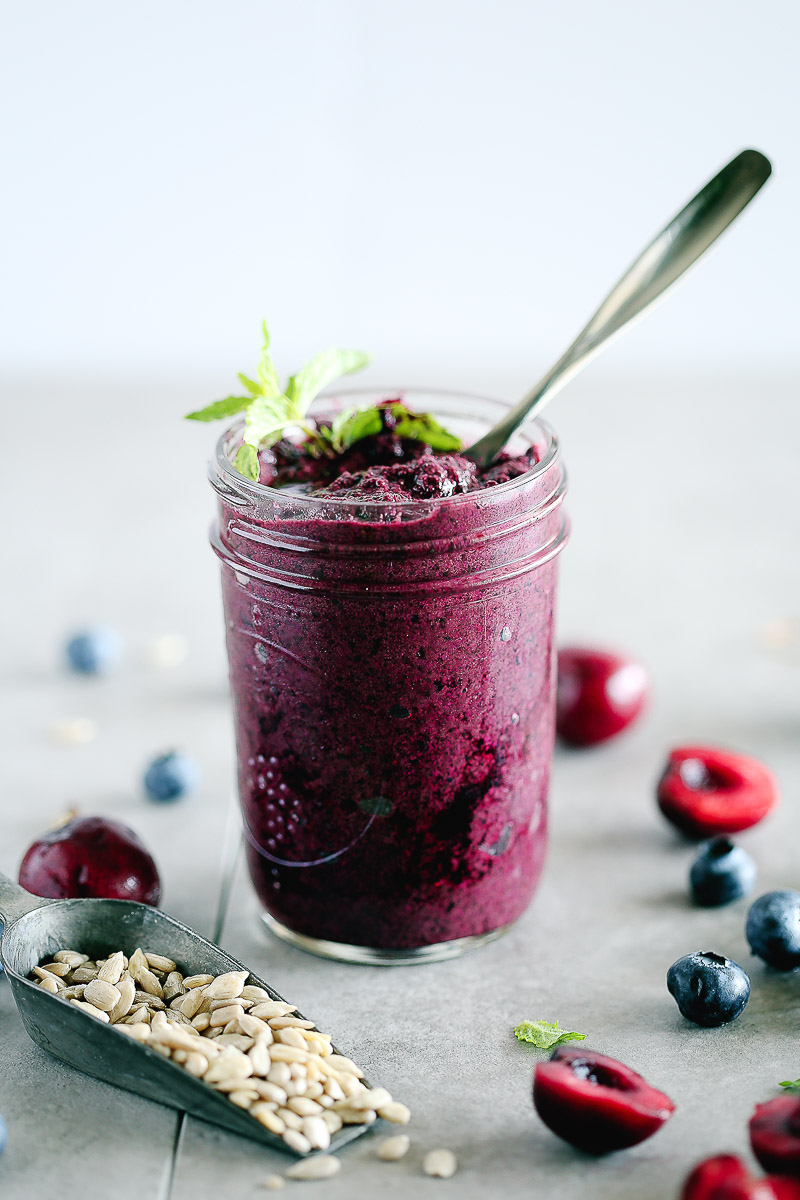 Blueberry Mint Grilled Cheese by Pasta-based. A small mason jar of pesto in a vibrant berry color. Ingredients like blueberries, cherries, mint, and sunflower seeds are scattered around.