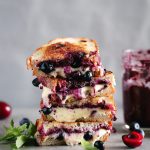 Blueberry Pesto Grilled Cheese by Pasta-based. A grilled cheese sandwich stacked high with gooey vegan mozzarella dripping down. Extra blueberries and cherries sprinkled around. Mint leaves, too.