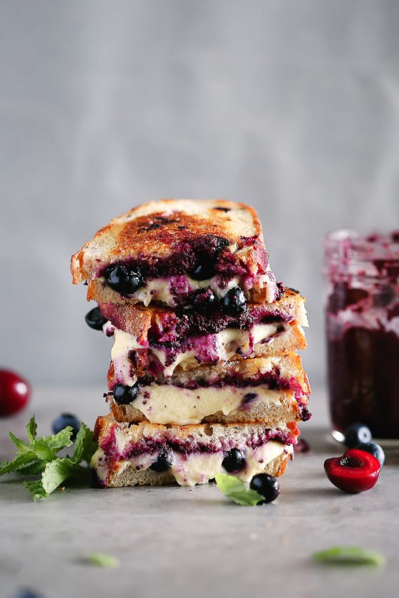 Blueberry Pesto Grilled Cheese by Pasta-based. A grilled cheese sandwich stacked high with gooey vegan mozzarella dripping down. Extra blueberries and cherries sprinkled around. Mint leaves, too.