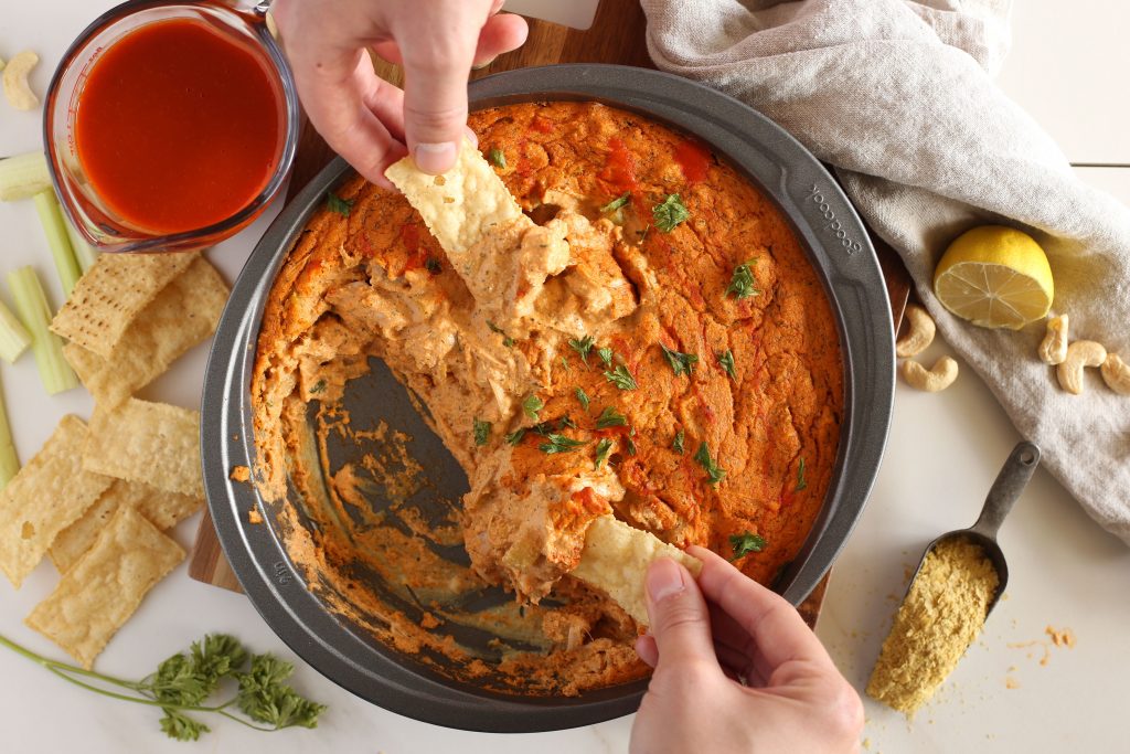 Overhead photo of buffalo jackfruit dip and two hands scooping some with chips.