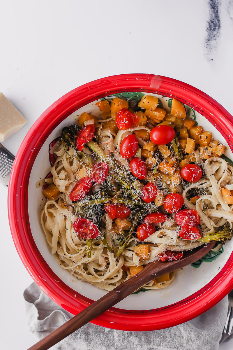 Creamy vegan pasta sauce with roasted butternut squash and broccolini. Recipe by Pasta-based. This photo shows a big bowl with ribbons of fettuccine, roasted butternut squash, broccolini and bright red grape tomatoes. Garnished with vegan parmesan cheese. 