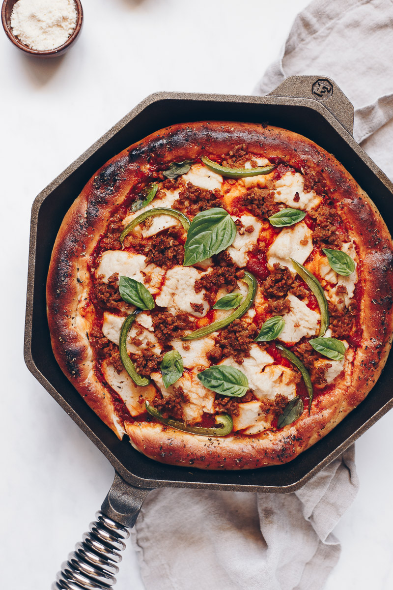 Vegan Deep Dish Pizza by Pasta-based. Thick crust pizza in a hot octagonal-shaped cast iron pan straight out of the oven. Thick golden brown crust, baked in a cast iron skillet with melty vegan mozzarella cheese and homemade plant-based sausage crumbles.