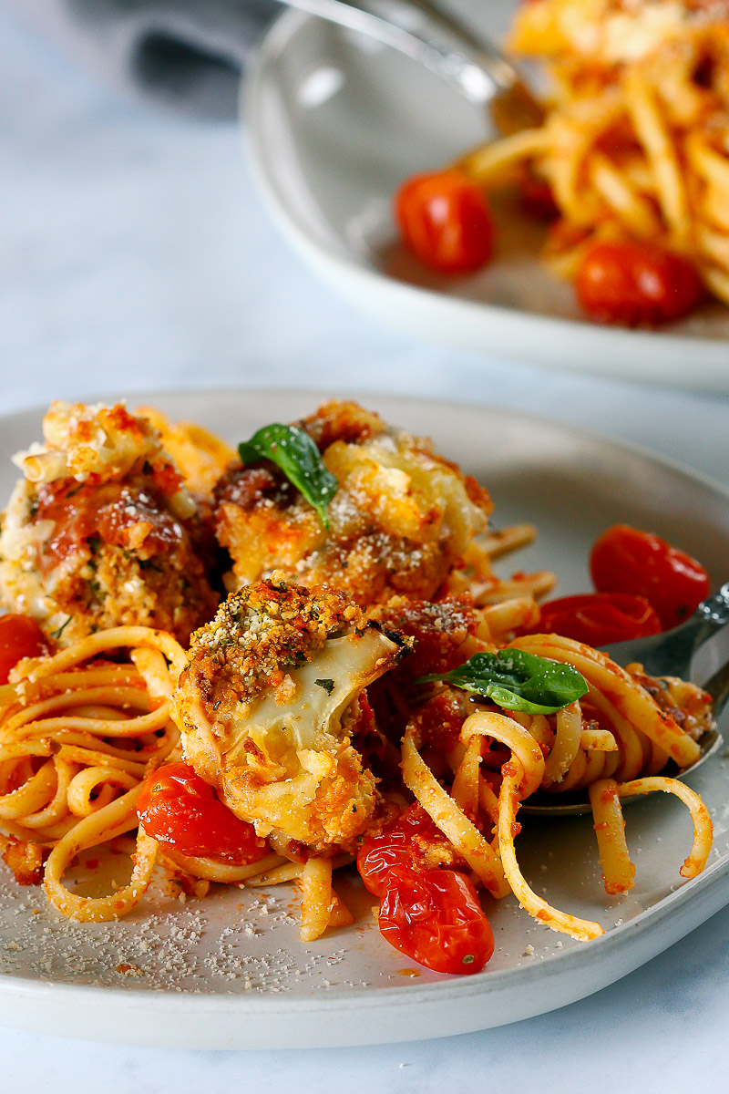 Vegan Cauliflower Parmesan by Pasta-based. A crispy, breaded cauliflower floret on top of a plate of linguine smothered in sauce with burst grape tomatoes.