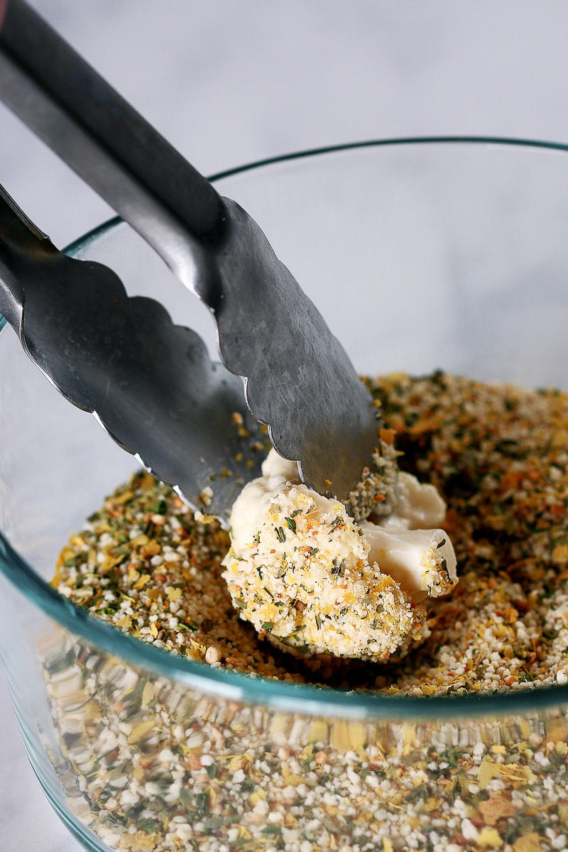 Vegan Cauliflower Parmesan by Pasta-based. A cauliflower floret being dipped into an Italian breadcrumb mixture using silver tongs.