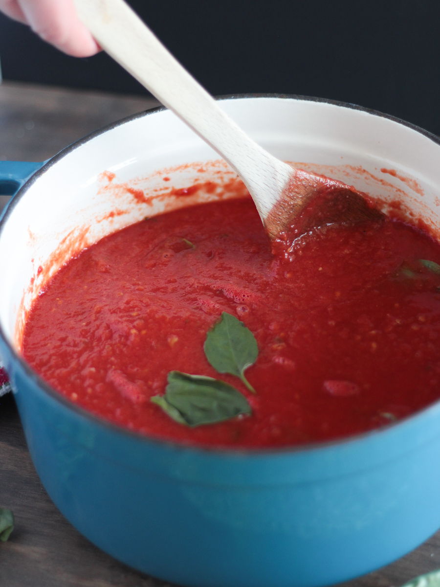 Side shot of tomato sauce in a big blue pot with basil floating on top.  Wooden spoon resting on pot.