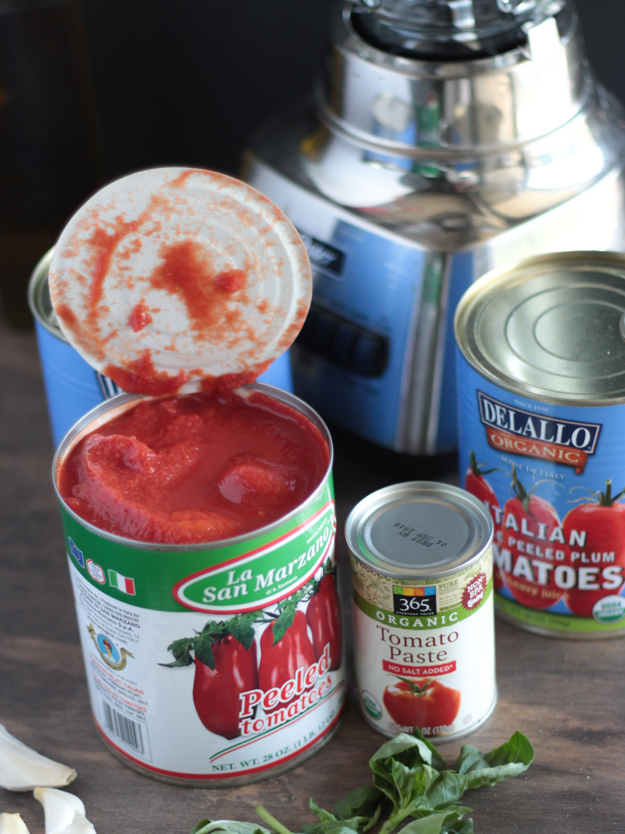 Tomato sauce ingredients picture.  San Marzano tomatoes in a can, tomato paste, garlic and a blender in the background.