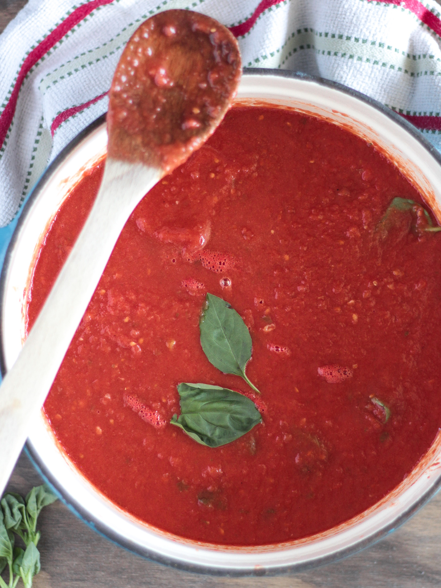 Overhead shot of sauce in a big blue pot with basil floating on top.  Wooden spoon resting on pot.