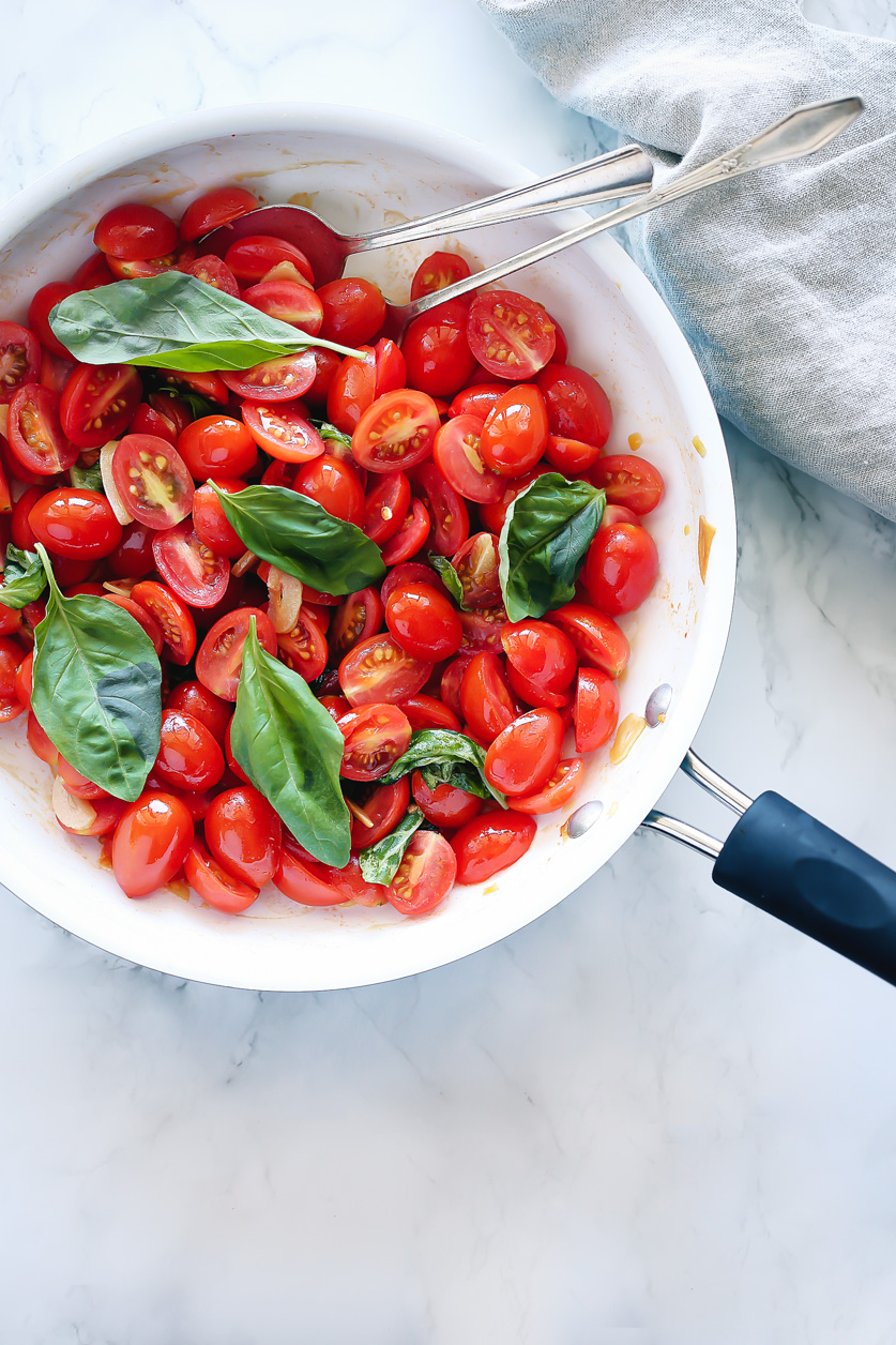 bright red grape tomatoes sliced in half and gathered in a white pan with fresh green basil before bursting open and cooking to form a light sauce.