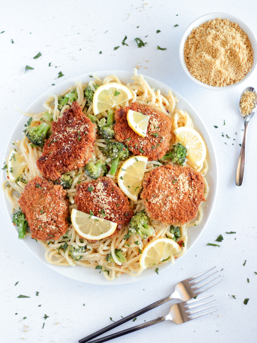 The Best chicken-style seitan cutlets on a white plate on top of spaghetti with broccoli and lemon slices.