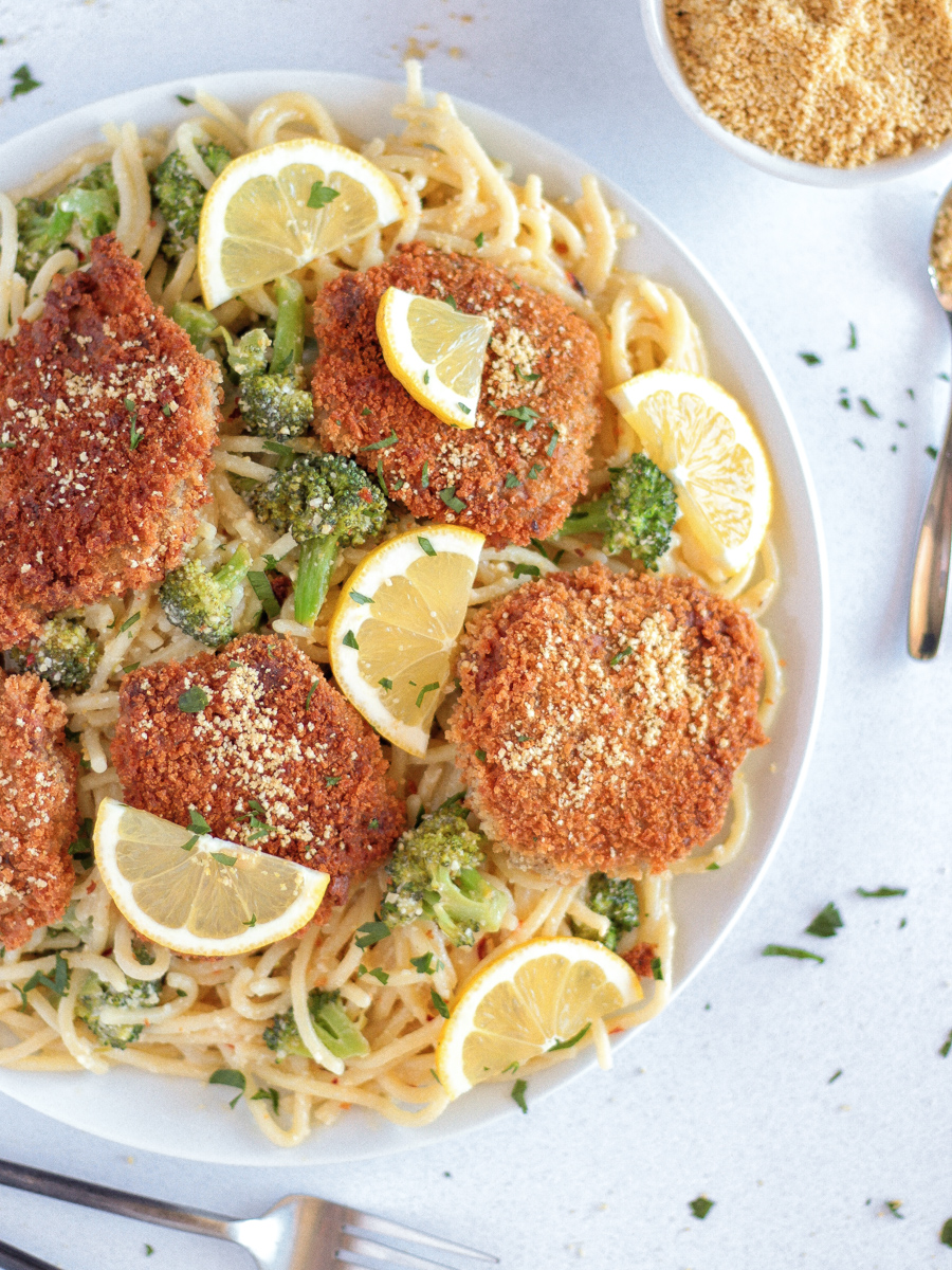 The Best chicken-style seitan cutlets on a white plate on top of spaghetti with broccoli and lemon slices.