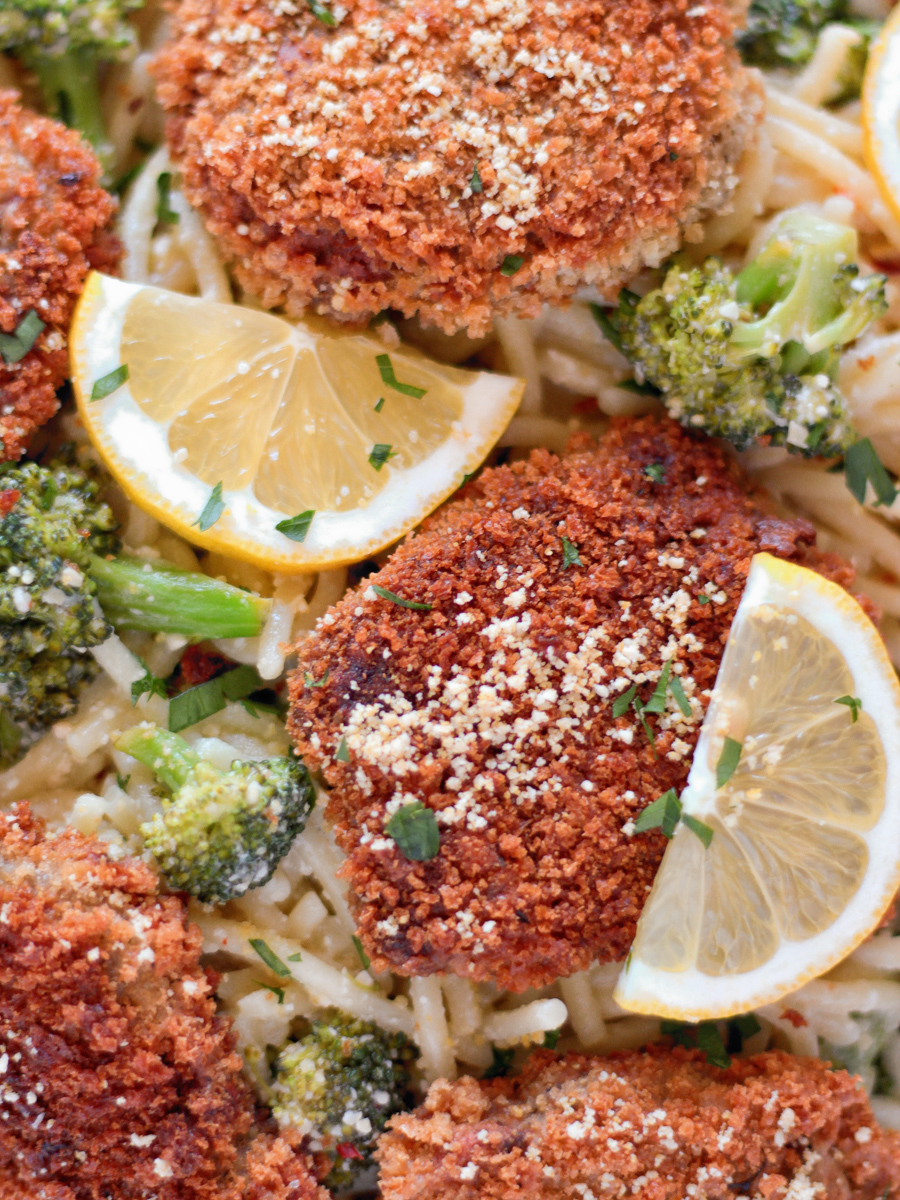 The Best chicken-style seitan cutlets on a white plate on top of spaghetti with broccoli and lemon slices.