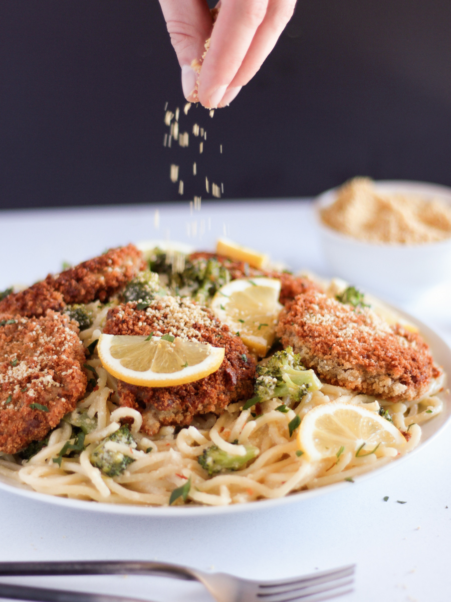 The Best chicken-style seitan cutlets on a white plate on top of spaghetti with broccoli and lemon slices.