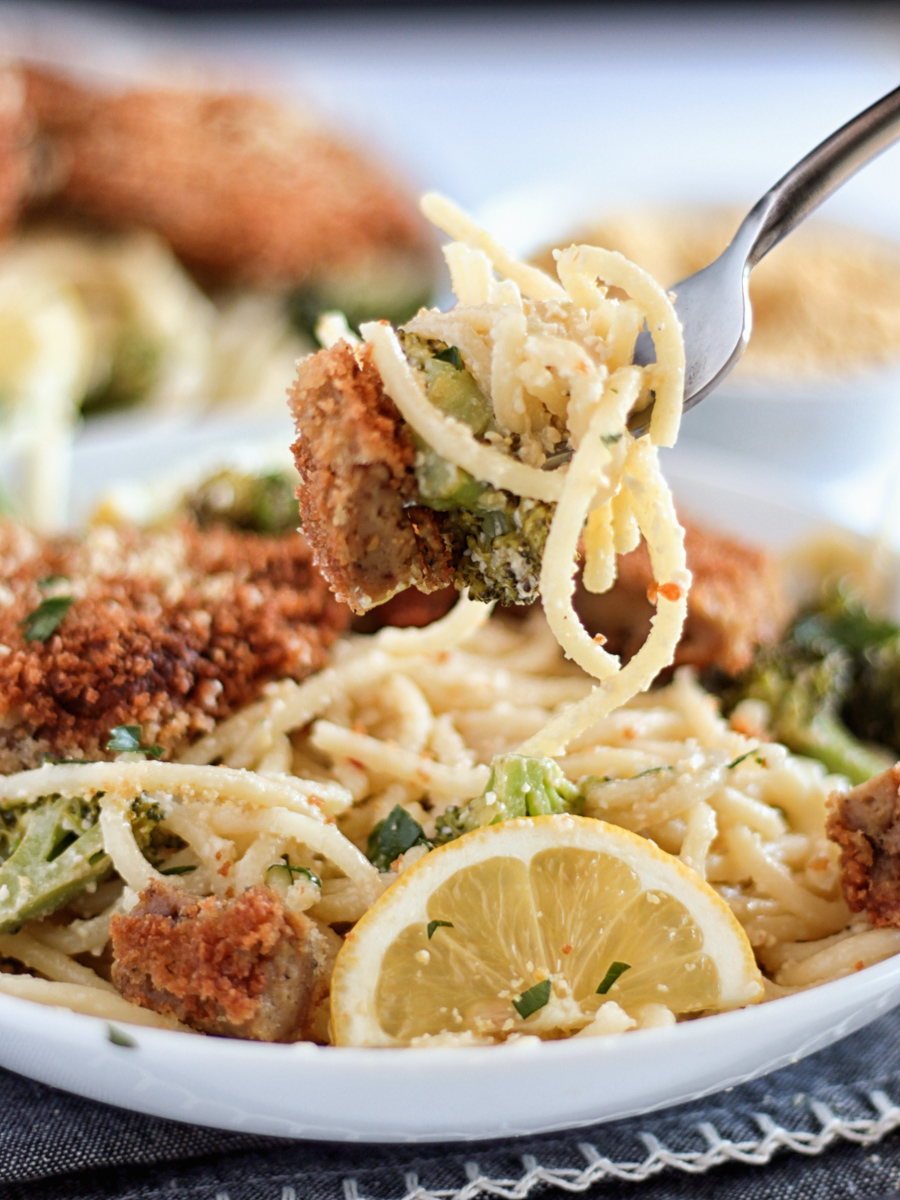 Fried vegan chicken cutlets on a white plate on top of spaghetti with broccoli and lemon slices.