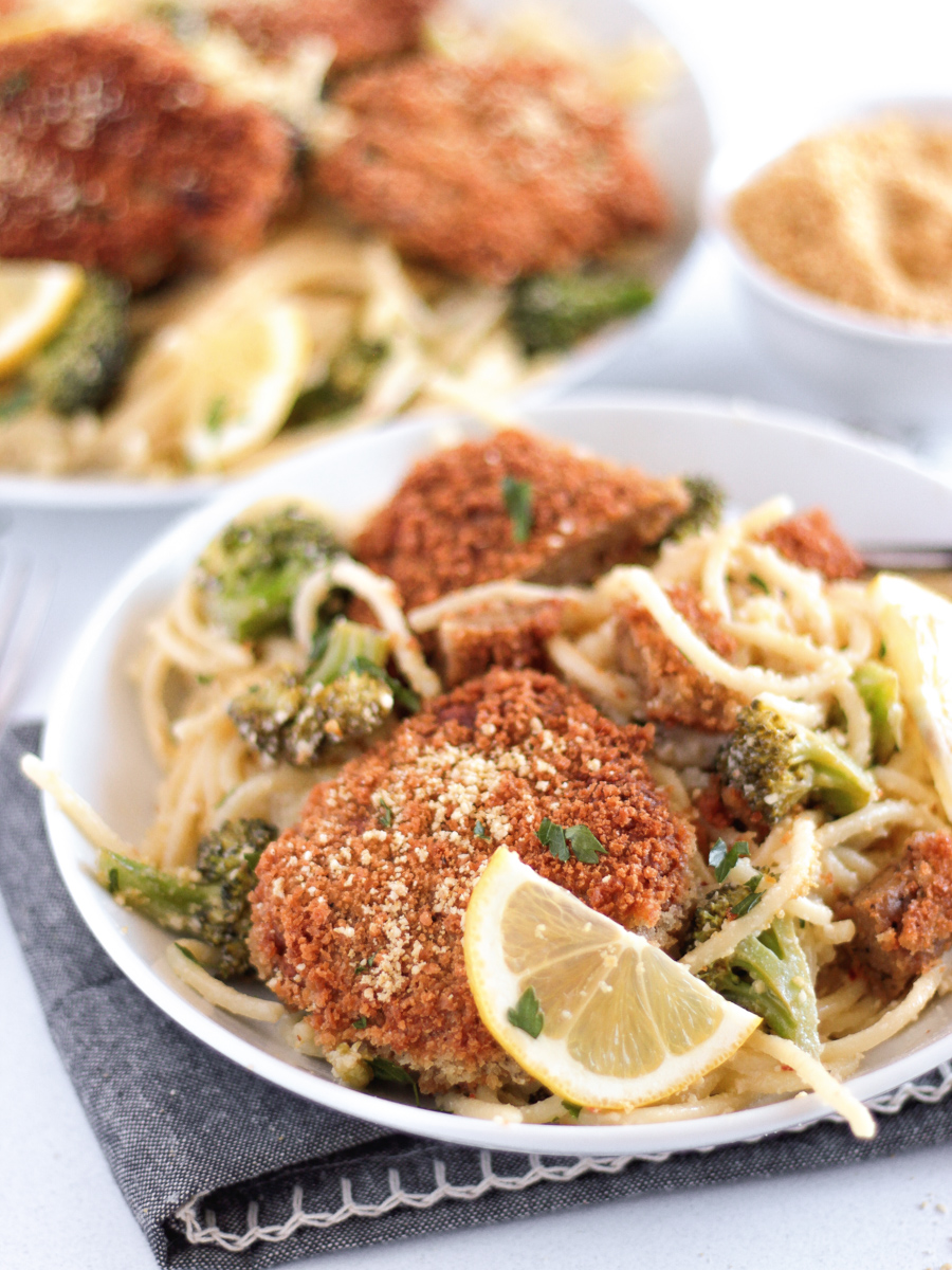 Fried vegan chicken cutlets on a white plate on top of spaghetti with broccoli and lemon slices.