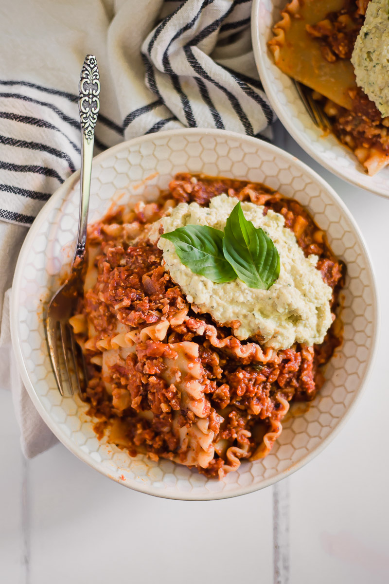 Vegan Lasagna Bowl by Pasta-based. All the ingredients of a lasagna but in a bowl! A large bowl stuffed with lasagna noodles, vegan ricotta cheese with two fresh basil leaves on top, and a vegan meat sauce made with cauliflower and walnuts.