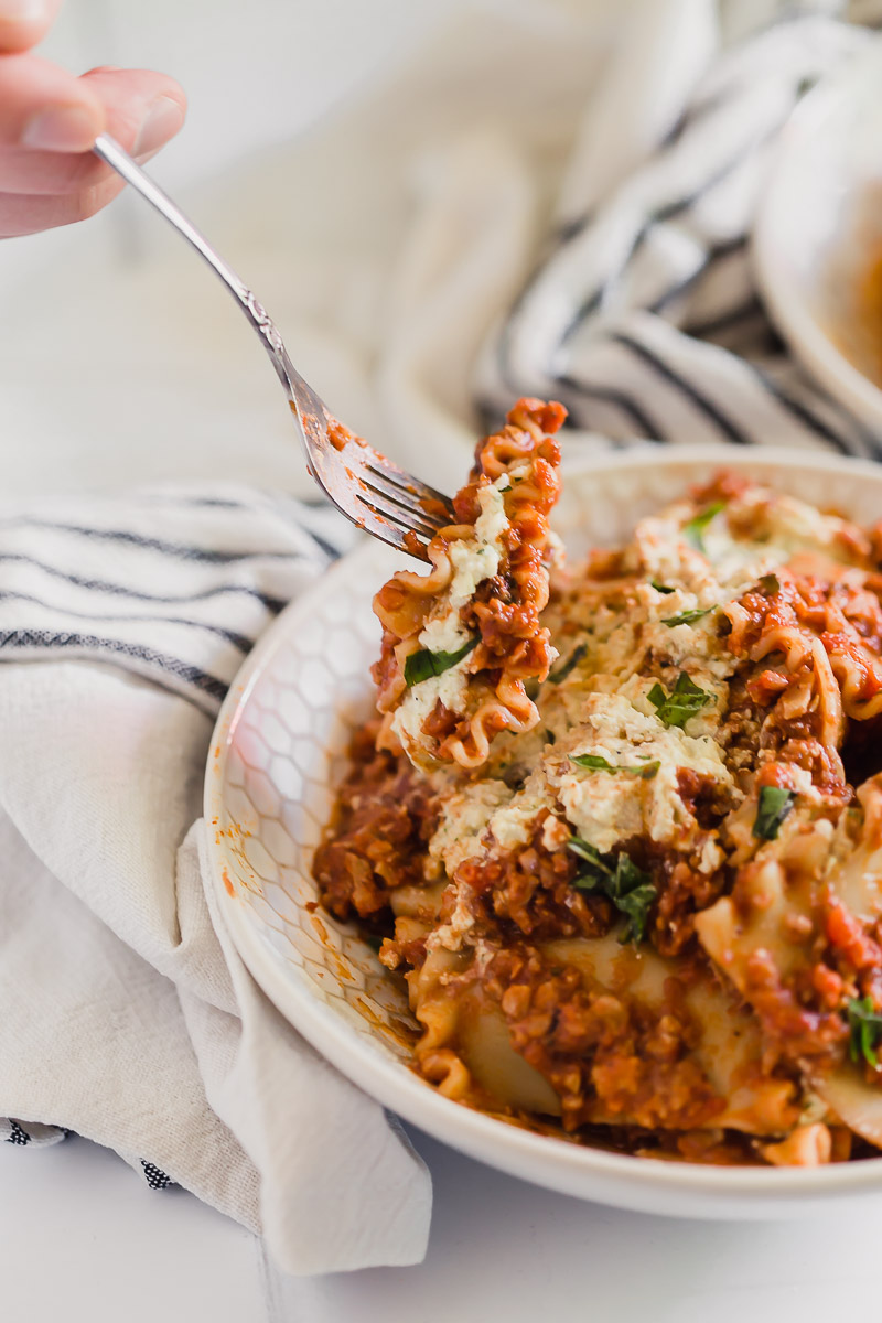 Vegan Lasagna Bowl by Pasta-based. All the ingredients of a lasagna but in a bowl! A close-up shot of a bowl filled with lasagna ingredients: wavy lasagne noodles, vegan meat sauce made with cauliflower and walnuts, and vegan ricotta cheese. Matt is holding a fork coming in from the left side and taking a forkful of the cheesy, saucy deliciousness.