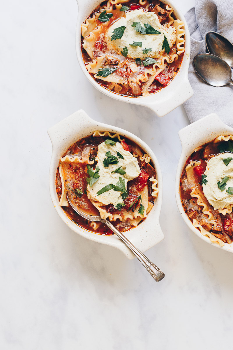 Vegan Lasagna Soup by Pasta-based. A bowl of brothy soup with sausage crumbles and lasagna noodles. There is a big dollop of vegan ricotta on top with parsley sprinkled on top of that. A piece of garlic bread accompanies the dish.