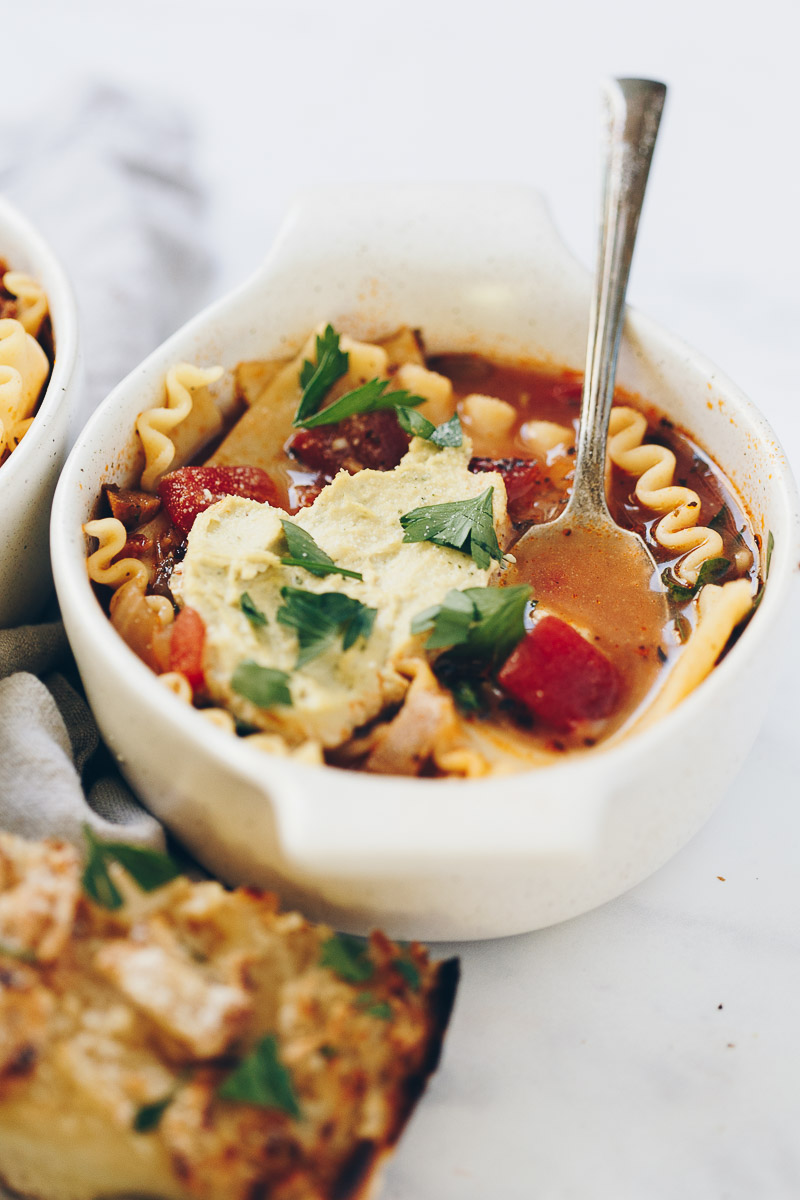 Vegan Lasagna Soup by Pasta-based. A bowl of brothy soup with sausage crumbles and lasagna noodles. There is a big dollop of vegan ricotta on top with parsley sprinkled on top of that. A piece of garlic bread accompanies the dish.