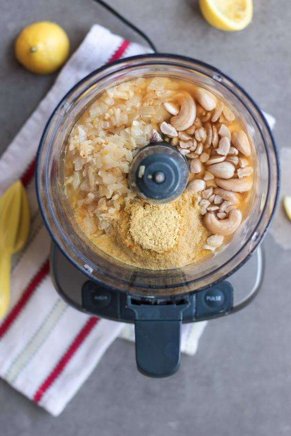 Easy White Wine Vegan Alfredo Sauce.  Overhead photo of open food processor with cashews, onions, nutritional yeast and lemon juice.  Lemon wedges scattered around the background.