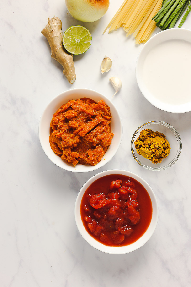 Vegan Pumpkin Curry Pasta by Pasta-based. Ingredients in bowls, scattered around on a marble table top. Ingredients in white bowls include diced tomatoes, pumpkin puree, and coconut cream. In a small clear bowl there is a couple tablespoons of yellow curry. On the side are garlic cloves, fresh ginger unpeeled, a lime sliced in half, green onions, a yellow onion, and dry fettuccine.