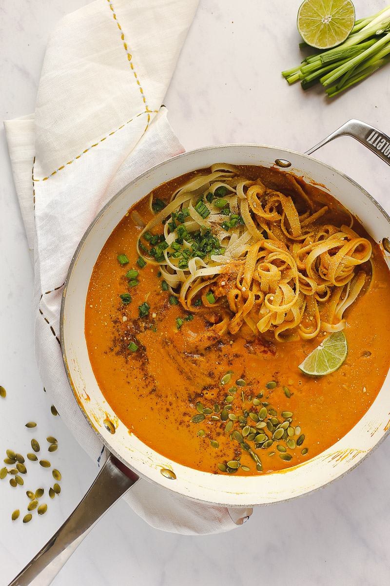 Vegan Pumpkin Curry Pasta by Pasta-based. A large pan with creamy, pumpkin curry pasta sauce with fettuccine, freshly chopped green onions, a sprinkle of nutmeg, roasted pepitas and a lime wedge. 