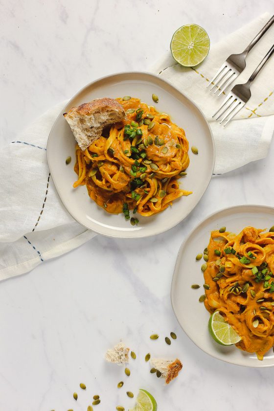 Vegan Pumpkin Curry Pasta by Pasta-based. Two white plates on a marble surface with a white embroidered dish towel underneath one of the plates. Two forks peak out the top-right of the frame beside a bright green lime sliced in half. Fettuccine smothered in a creamy, pumpkin curry pasta sauce is piled in the middle of both plates. Both plates have garnishes like roasted pepitas, freshly chopped green onion, lime wedges, and kalamata-olive garlic bread.