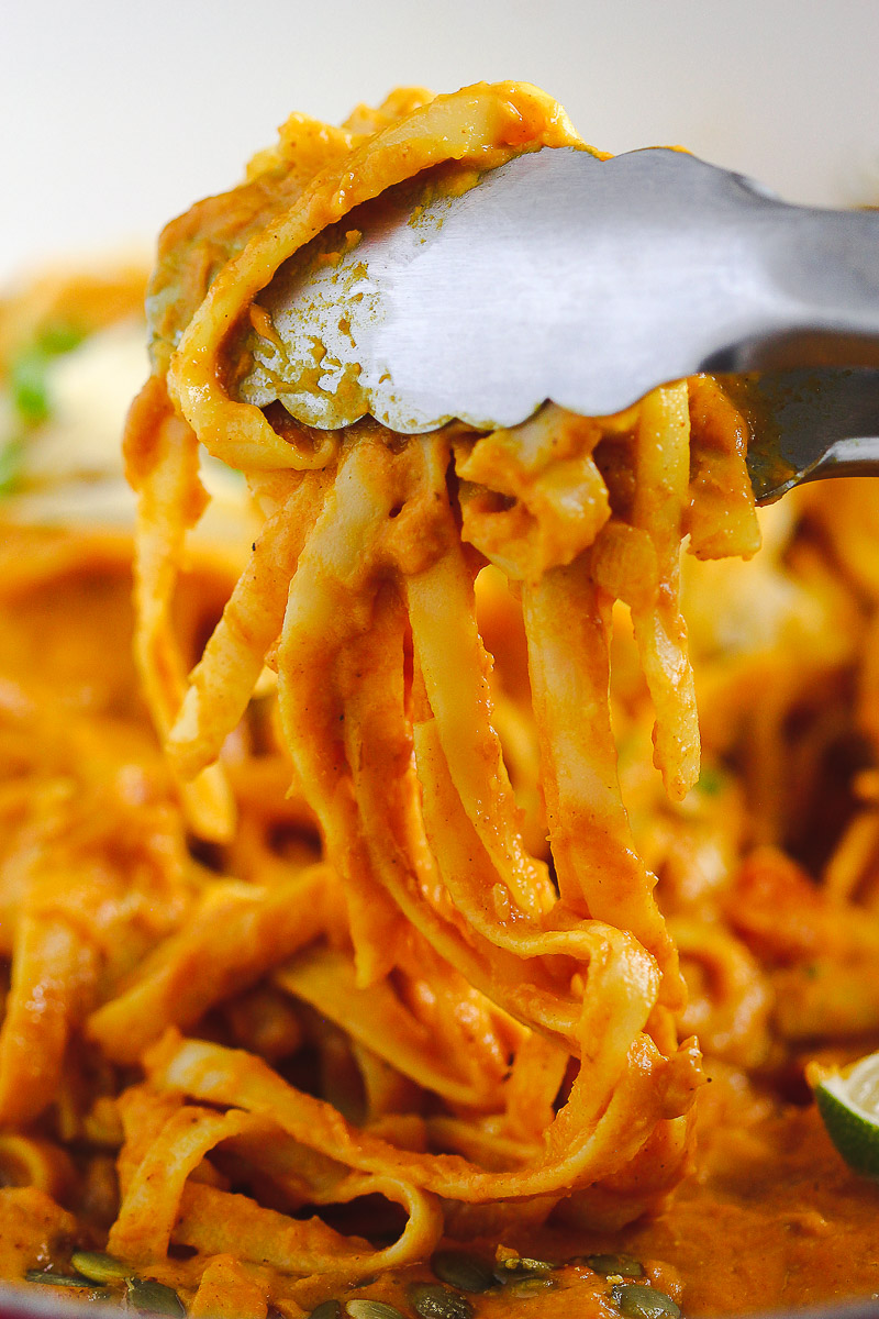 Vegan Pumpkin Curry Pasta by Pasta-based. A close-up shot of a pair of tongs mixing cooked fettuccine with the creamy, luscious, pumpkin-coconut curry pasta sauce. The pasta sauce is a bright pumpkin orange and looks like it's smothered in the sauce and extra creamy and delicious.