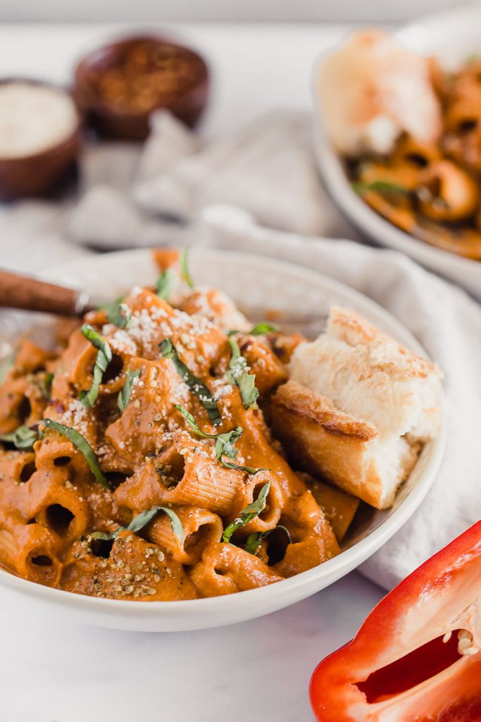 Vegan Roasted Red Pepper Pasta Sauce by Pasta-based. A bowl of rigatoni smothered with creamy roasted red pepper sauce and a piece of crusty Italian bread on the side. Ribbons of basil leaves sprinkled on top and a fork with a wooden handle rested on top. This pasta is ready to be devoured!