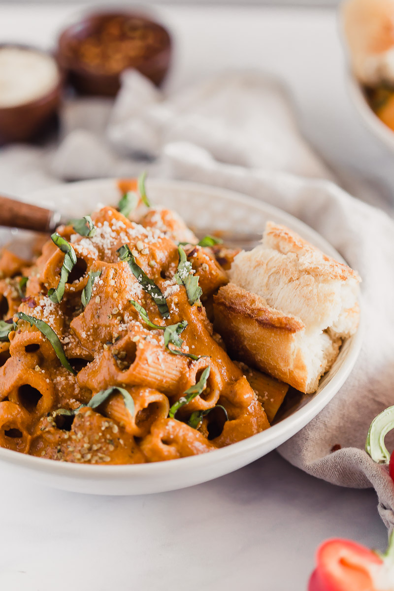 Vegan Roasted Red Pepper Pasta Sauce by Pasta-based. A bowl of rigatoni smothered with creamy roasted red pepper sauce and a piece of crusty Italian bread on the side.