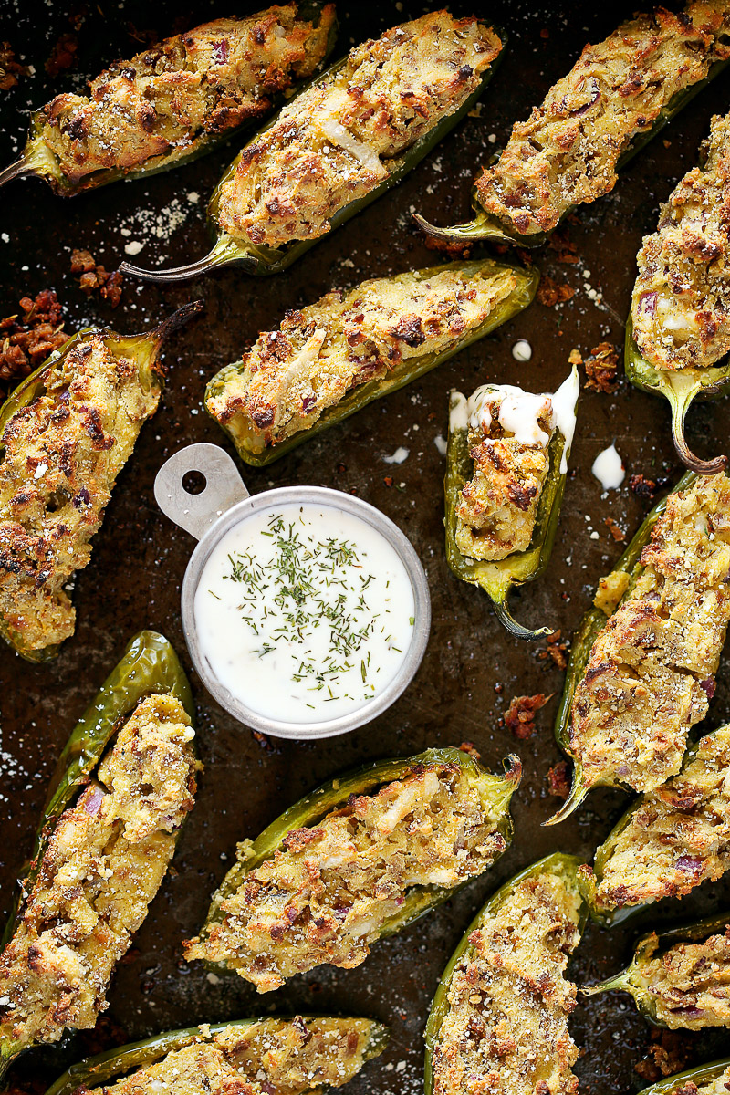 Vegan Jalapeno Poppers by Pasta-based. Beautiful, bursting, crispy, jalapeno poppers stuffed with vegan ricotta and sausage crumbles sprawled out on a dark grey baking sheet. Small bowl of vegan ranch with fresh dill and a jalapeno drenched in ranch with a bite taken out.