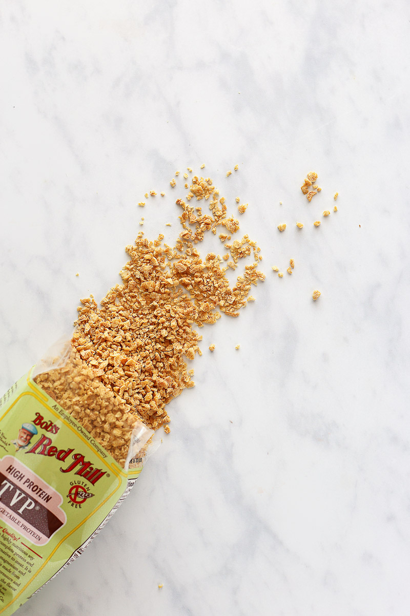 Vegan Italian Sausage Crumbles by Pasta-based. Textured Vegetable Protein (TVP) from Bob's Red Mill spills out of the bag onto a clean marble countertop. The TVP is a yellowy-brown and looks crunchy in texture.