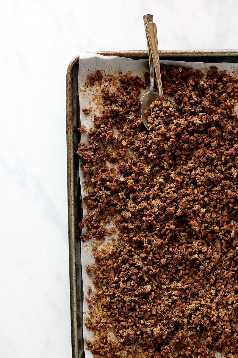 Vegan Italian Sausage Crumbles by Pasta-based. A metal baking pan on a white marble countertop with vegan italian sausage crumbles scattered onto a piece of parchment paper on the pan. To vintage spoons lean against the top of the baking pan with vegan italian sausage crumbles scooped into one of the spoons.