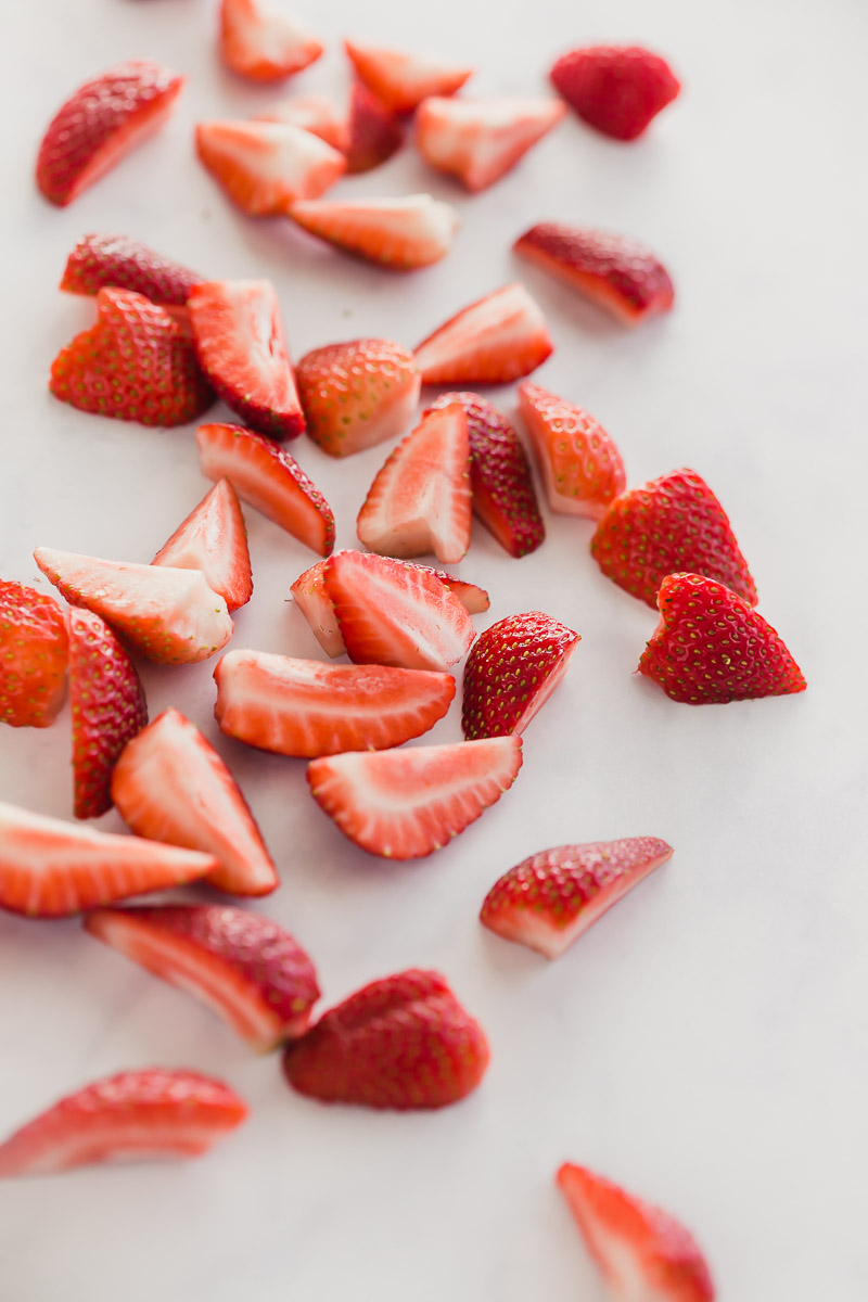 Vegan Strawberry Buttercream Cupcakes by Pasta-based. Bright red, juicy, sliced strawberries scattered on a light grey surface.