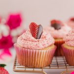 Vegan Strawberry Buttercream Cupcakes by Pasta-based. The cutest strawberry cut into a heart shape and dipped halfway in chocolate. Lined up on a cooling rack with other strawberry buttercream cupcakes and a dozen pink roses in the background. Perfect for Valentine's Day or any special occasion.