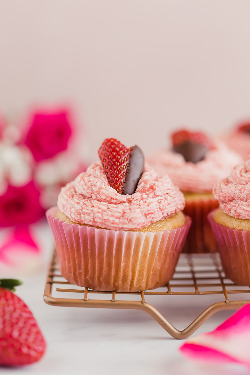 Vegan Strawberry Buttercream Cupcakes by Pasta-based. The cutest strawberry cut into a heart shape and dipped halfway in chocolate. Lined up on a cooling rack with other strawberry buttercream cupcakes and a dozen pink roses in the background. Perfect for Valentines Day or any special occasion.