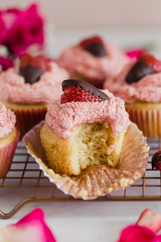 Vegan Strawberry Buttercream Cupcakes by Pasta-based. Fresh strawberries whipped with vegan butter on top of vanilla bean cupcakes. Topped with chocolate covered strawberries. This photo shows that someone took a bite out of the cupcake, the texture is airy light and fluffy. The buttercream is fresh and decadent. Rose petals are scattered around.