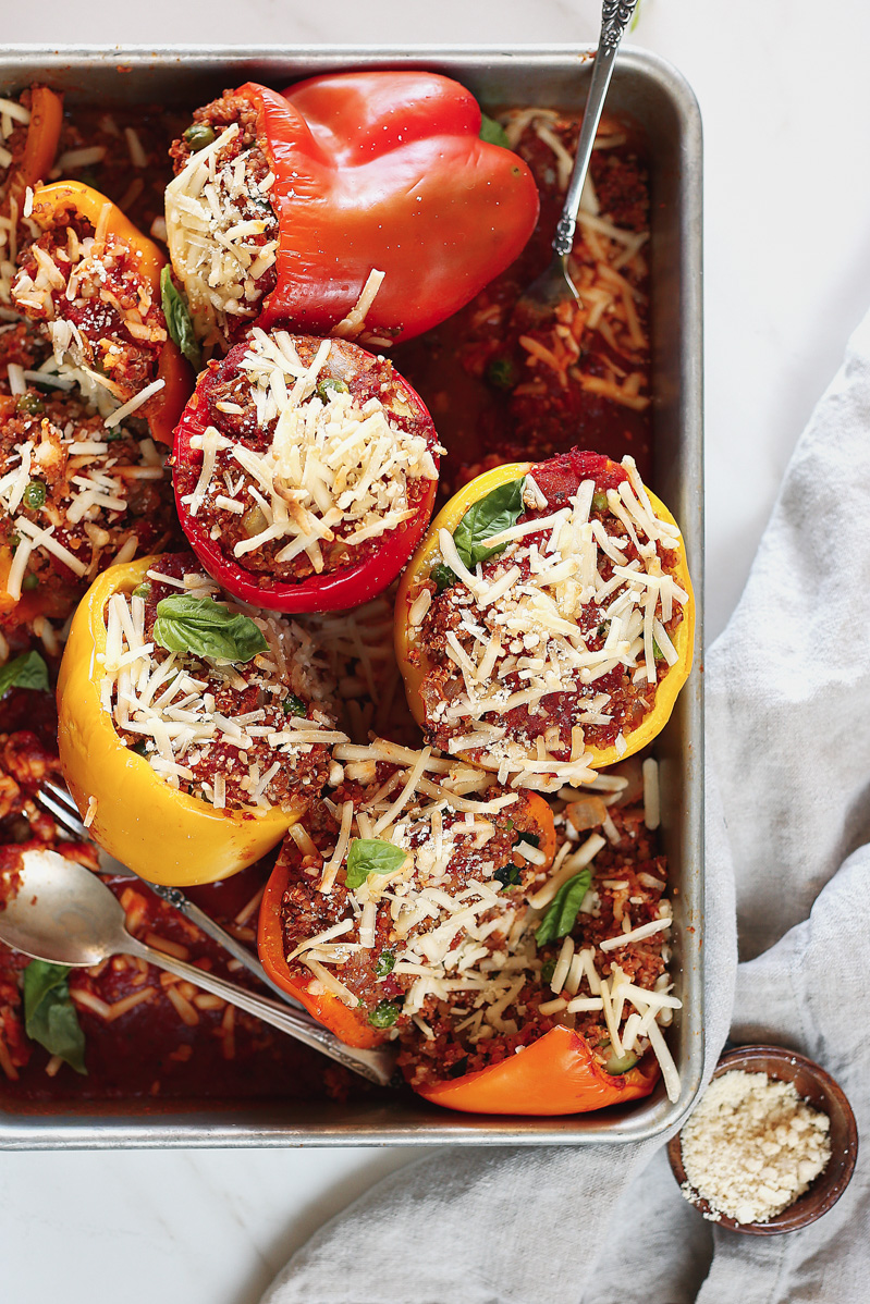 vegan stuffed peppers by pasta-based. vegan quinoa stuffed bell peppers in red, orange, and yellow gathered in a large silver pan with vegan mozzarella cheese and extra marinara sauce on top.