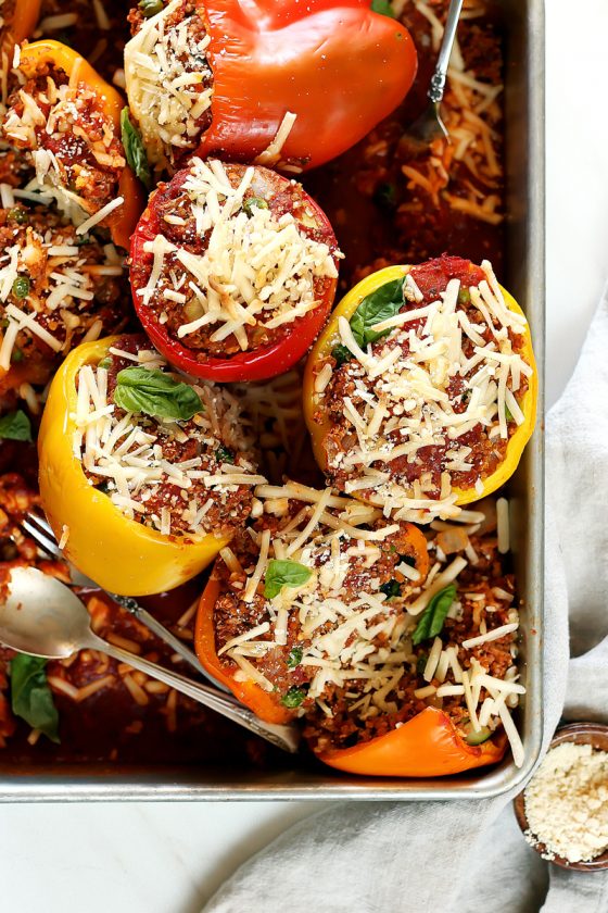 vegan stuffed peppers by pasta-based. vegan quinoa stuffed bell peppers in red, orange, and yellow gathered in a large silver pan with vegan mozzarella cheese and extra marinara sauce on top.