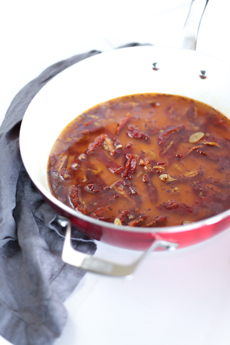 A photo of sun dried tomatoes in a white wine, vegetable broth sauce in a large red pan on top of a dark grey cloth.