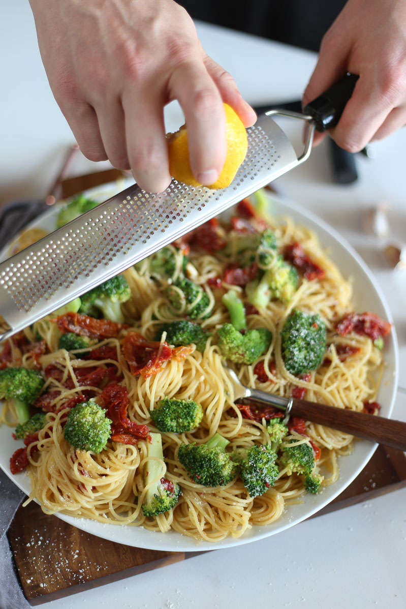 A large white plate filled to the top with sun dried tomato pasta and broccoli florets. Someone is holding a zester and zesting lemon on top of the pasta.