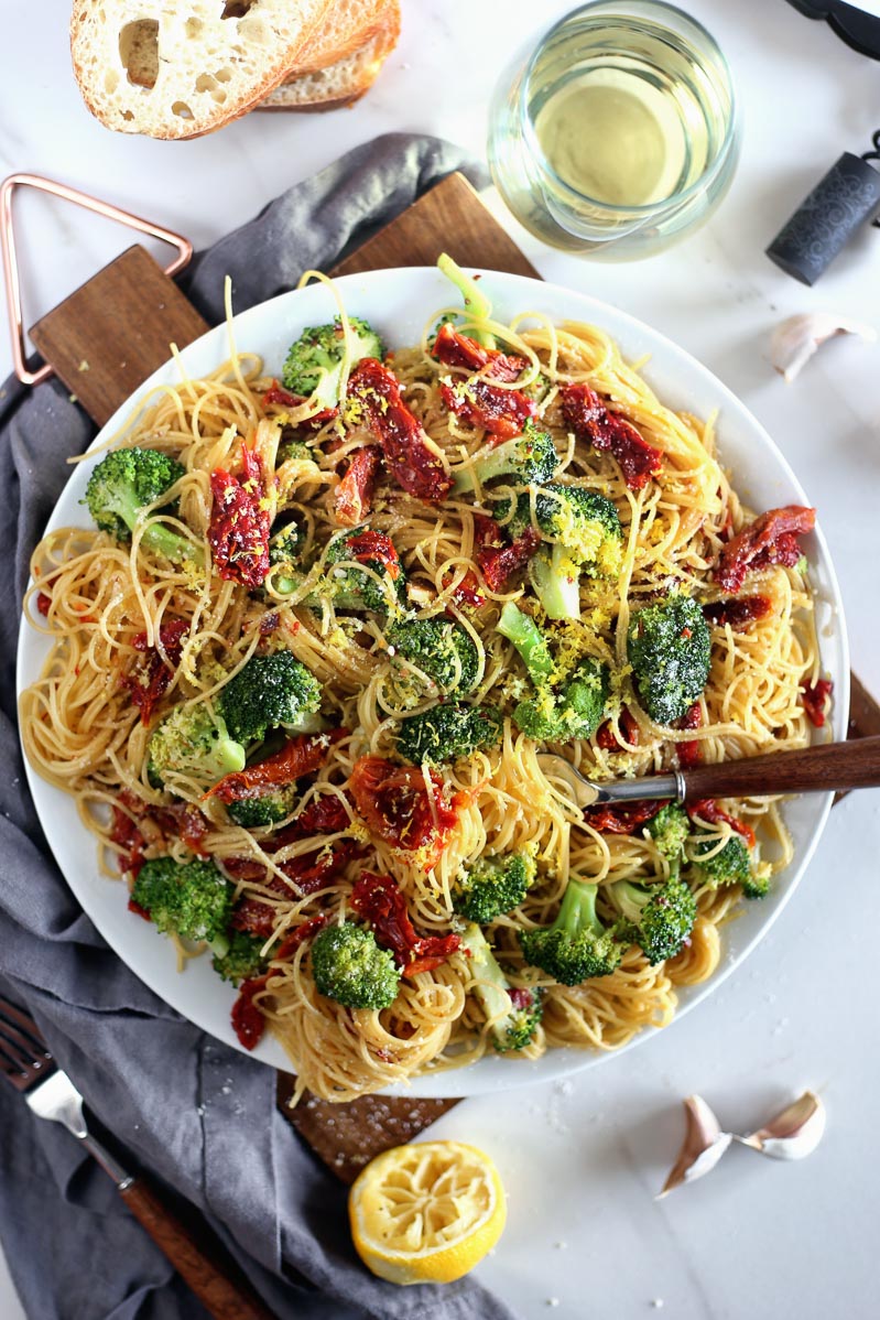 A large plate filled with angel hair pasta in a sun dried tomato, white wine, vegetable broth sauce. Also on the table is a glass of white wine and a wine cork, slices of Italian bread, a squeezed lemon, a fork with a wooden handle, cloves of garlic, and a dark grey cloth.