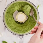 Nut free vegan pesto by Pasta-based. An overhead shot of a food processor with creamy pesto inside. A hand comes into the frame and scoops some of the pesto with a spoon.