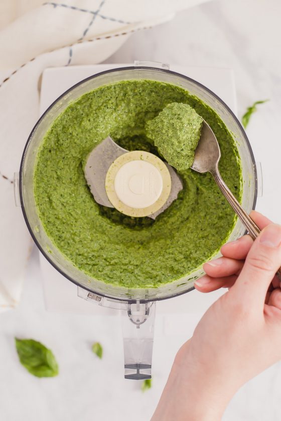 Nut free vegan pesto by Pasta-based. An overhead shot of a food processor with creamy pesto inside. A hand comes into the frame and scoops some of the pesto with a spoon.