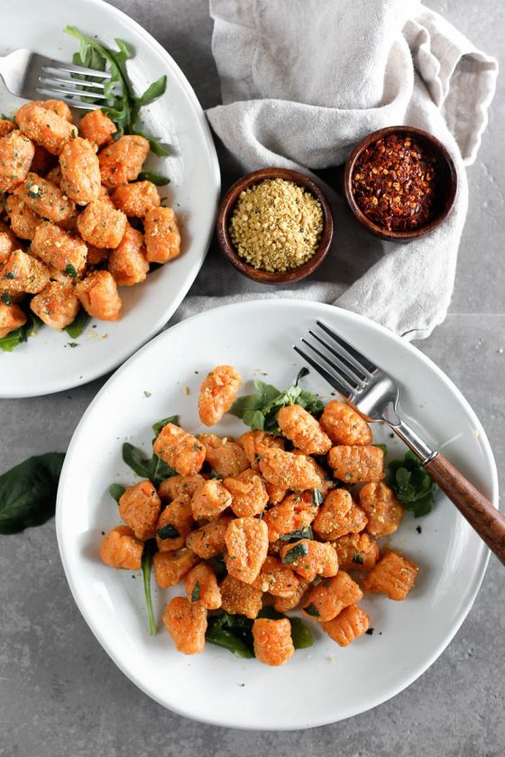 Vegan Sweet Potato Gnocchi by Pasta-based. Overhead photo of two white plates with cooked sweet potato gnocchi. Two small wooden bowls filled with cashew parmesan and red pepper flakes on the side.