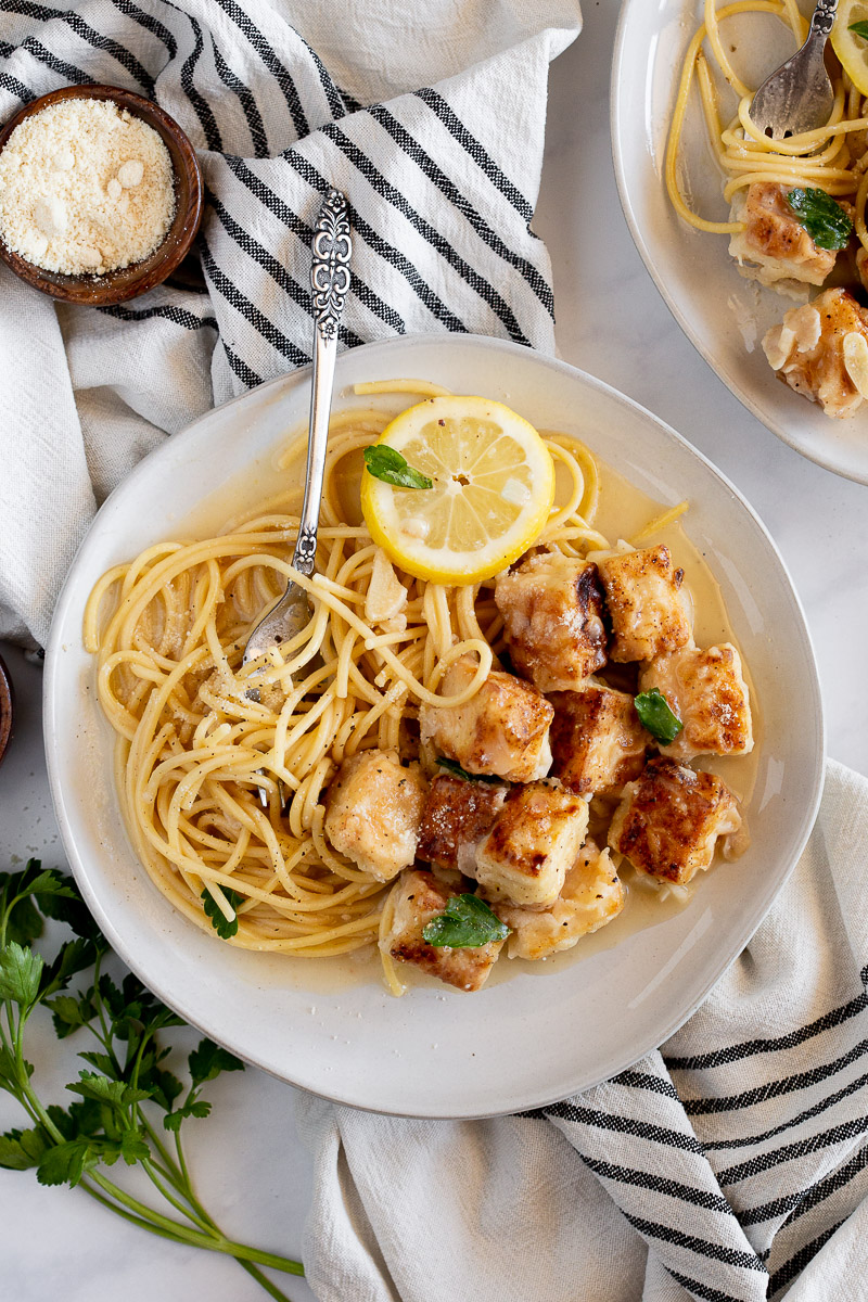 An overhead view of two white bowls on a marble table. One bowl rests on top of a linen cloth. Inside the bowl is Vegan Chicken Francese made of browned tofu bites and a lemony-butter sauce. Also in the bowl is garlicky spaghetti with a garnish of vegan parmesan cheese and fresh parsley.