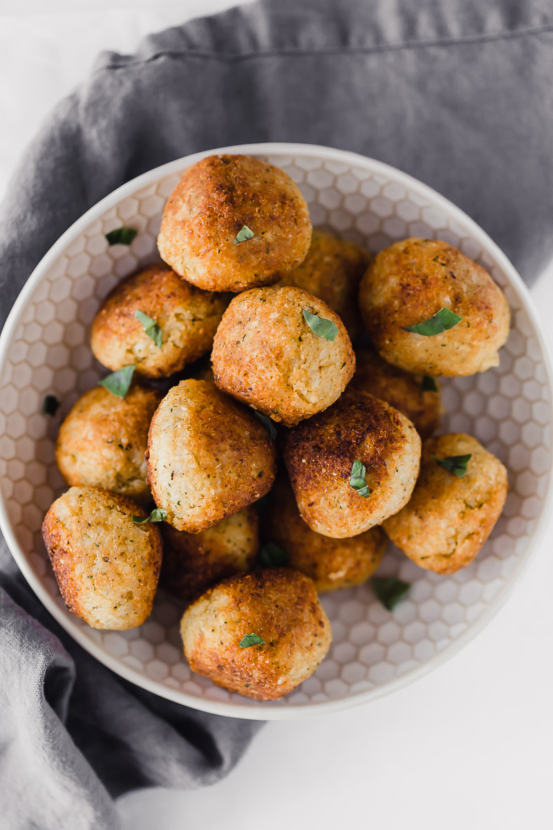 Golden brown vegan meatballs piled up in a white textured bowl. Laying on top of a dark grey linen towel.