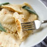 Vegan Butternut Squash Ravioli by Pasta-based. A butternut squash ravioli cut in half and on a fork, resting on top of the other raviolis in a dish, showing the delicious filling stuffed inside.