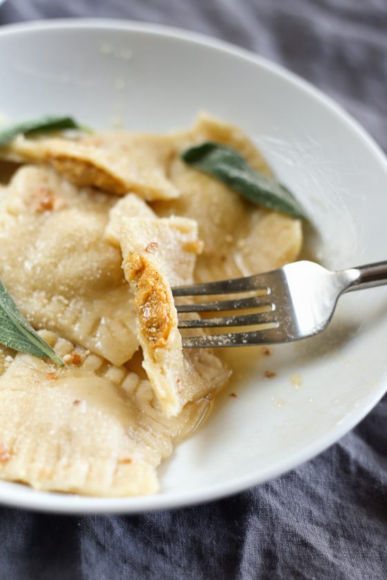 Vegan Butternut Squash Ravioli by Pasta-based. A butternut squash ravioli cut in half and on a fork, resting on top of the other raviolis in a dish, showing the delicious filling stuffed inside.