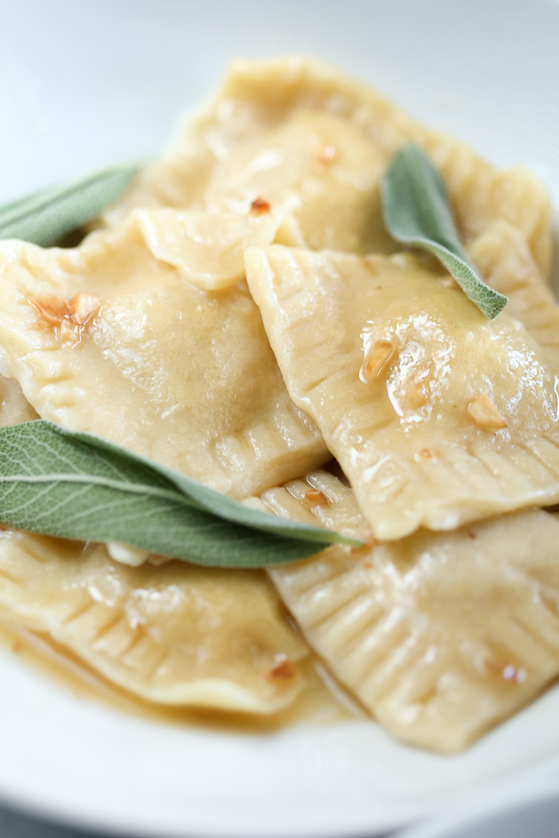 Vegan Butternut Squash Ravioli by Pasta-based. Finished and cooked ravioli nestled together on a white plate covered in a sage, garlic, brown-butter sauce with fresh sage leaves for garnish.